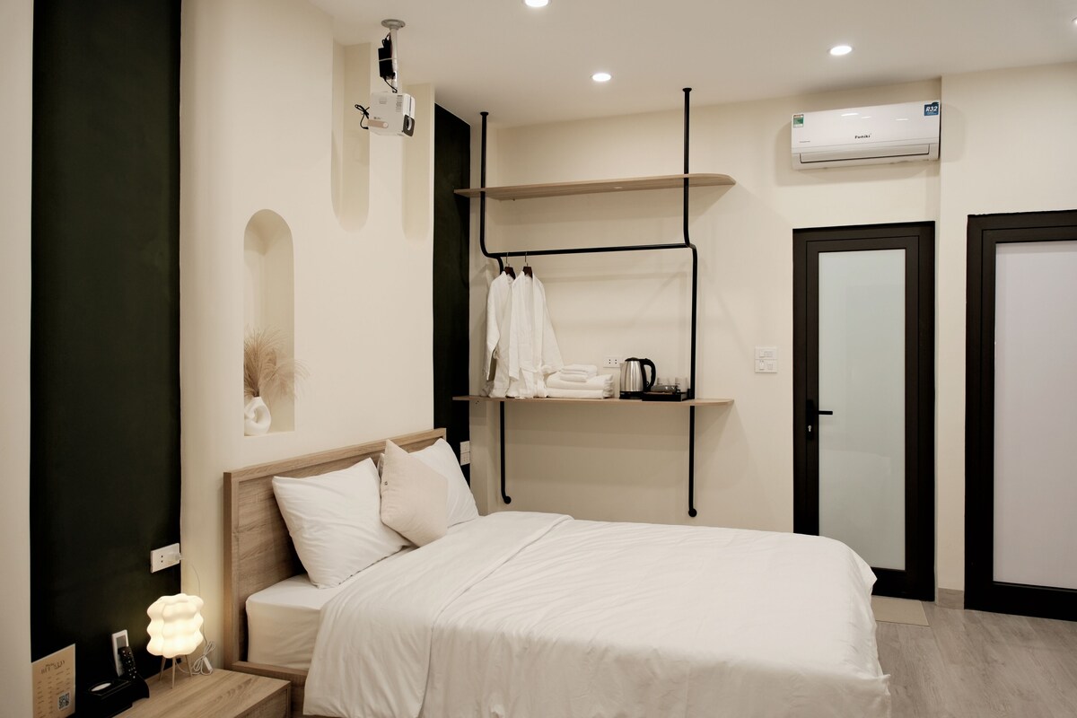 Una habitación de hotel moderna con cama, mesita de noche, repisa en la pared con toallas, aire acondicionado en la pared y una puerta.
