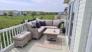 Ein überdachter Sitzbereich im Freien mit einer Couch und einem Couchtisch, einer weißen Geländer und Blick auf ein Rasenfeld.