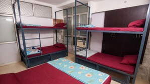 A dormitory room with bunk beds, a small table with a floral tablecloth, and a window with a metal screen. - Vrindavan Bunk Hostel by Shiva - 5min Walk from Prem Mandir (Vrindavan)
