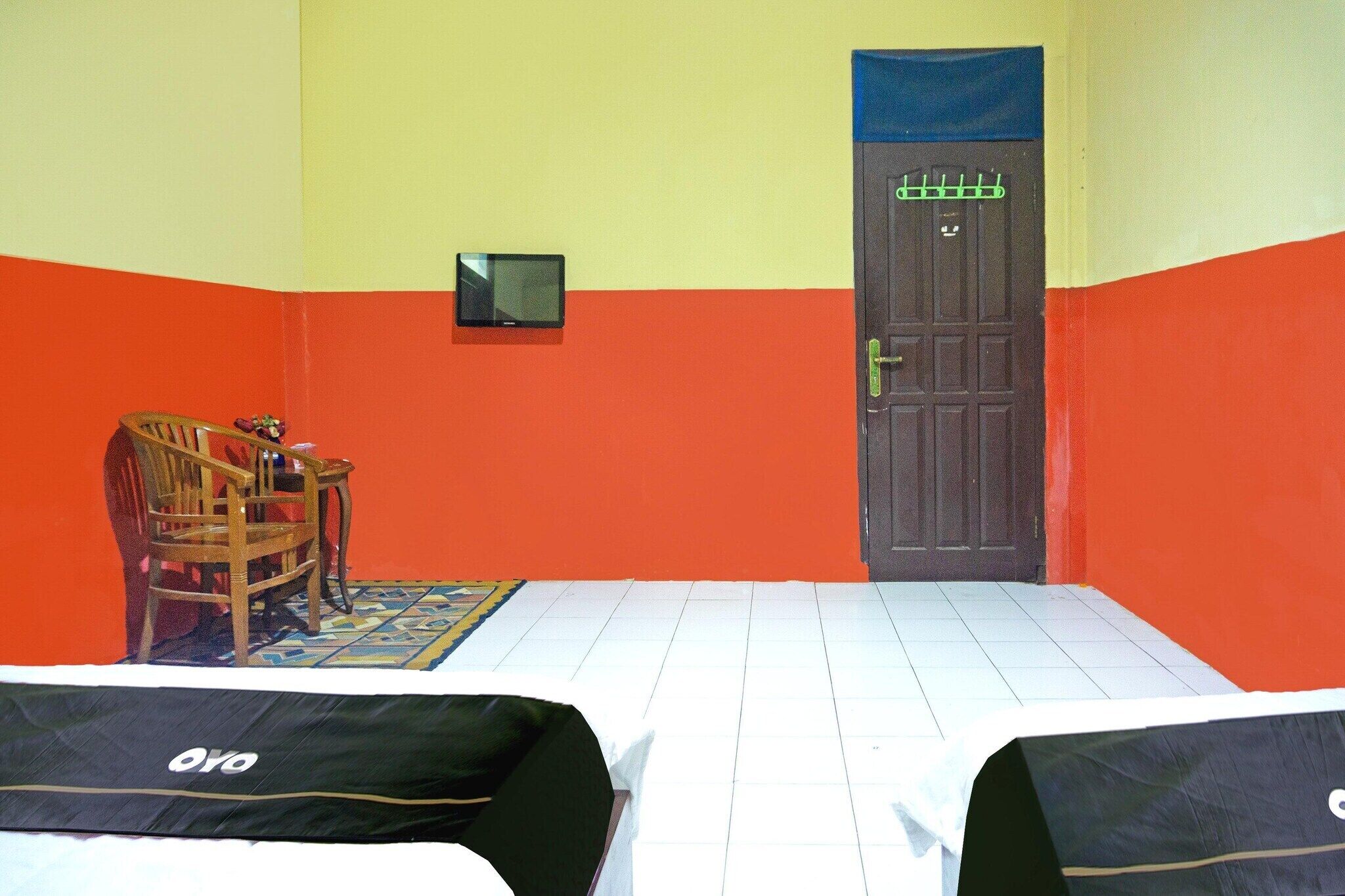 Ein Zimmer mit zwei Betten, einem Sessel, einem kleinen Tisch, einer Tür und einem an der Wand befestigten Fernseher.