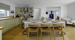 A modern kitchen with wooden cabinets, a dining table set for four, and a view into the living area. - Hampton Coast (Abersoch)