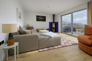 A modern living room with a sofa, a table, a lamp, a fireplace, and a large window. - Hampton Coast (Abersoch)