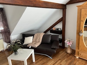 Ein Wohnzimmer mit einem dunkelbraunen Sofa, einem weißen Tisch mit Pflanze, Holzbalken, einem Holzschrank und einem kleinen Regal mit Radio und Dekorationsobjekt.