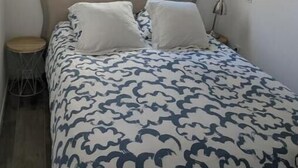 Un dormitorio con una cama, un taburete pequeño y redondo, una ventana y una lámpara.