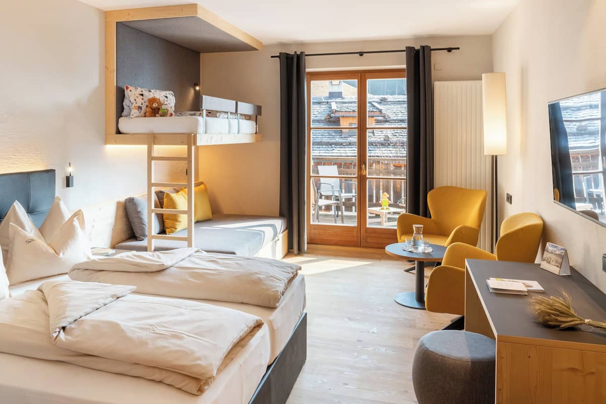 A hotel room with a bunk bed, a desk with a chair, a small table with a lamp, and a view of the outside.