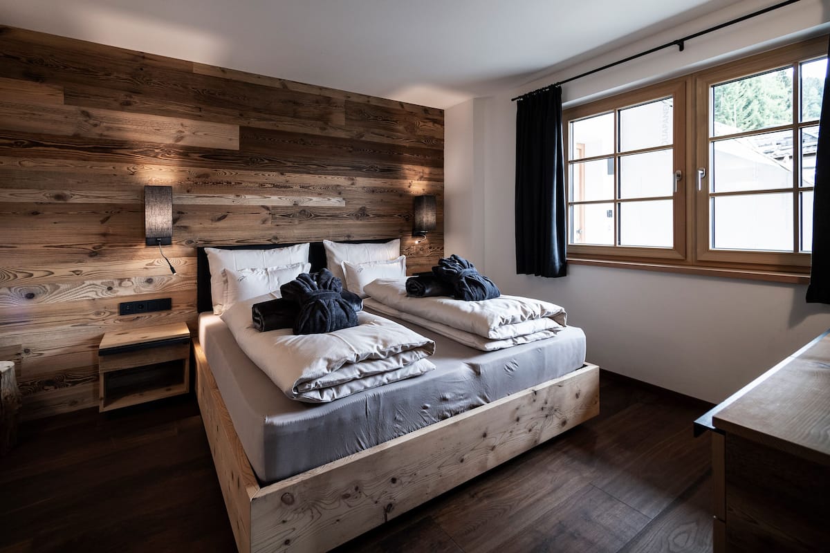 A bedroom with a wooden headboard, a bed with pillows and blankets, bedside tables, a nightstand, and a window with curtains.