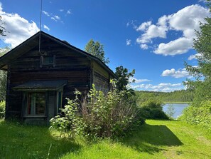 Maalaishenkinen, puinen mökki, jossa on kalteva katto, pieni kuisti ja ikkuna.