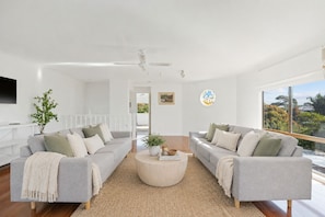 Ein modernes Wohnzimmer mit zwei grauen Sofas, einem runden Couchtisch, einem Deckenventilator, einem großen Fenster und einer weißen Wand mit einem gerahmten Bild und einem Buntglasfenster.