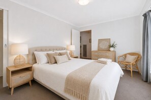 A bedroom with a large bed, two bedside tables, a wooden dresser, a wicker chair, and a mirror on the wall. - Escape Away by Ready Set Stay (Cowes)