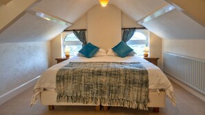 A bedroom with a sloped ceiling, a bed with a green blanket, two pillows, a nightstand with a lamp, and a window with curtains. - St David's Country House (Woodford)