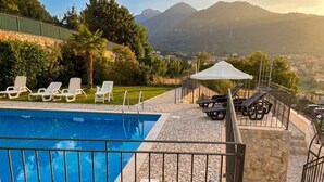 Ein Pool mit einer Terrasse aus Steinplatten, weißen Liegestühlen und Blick auf Berge.