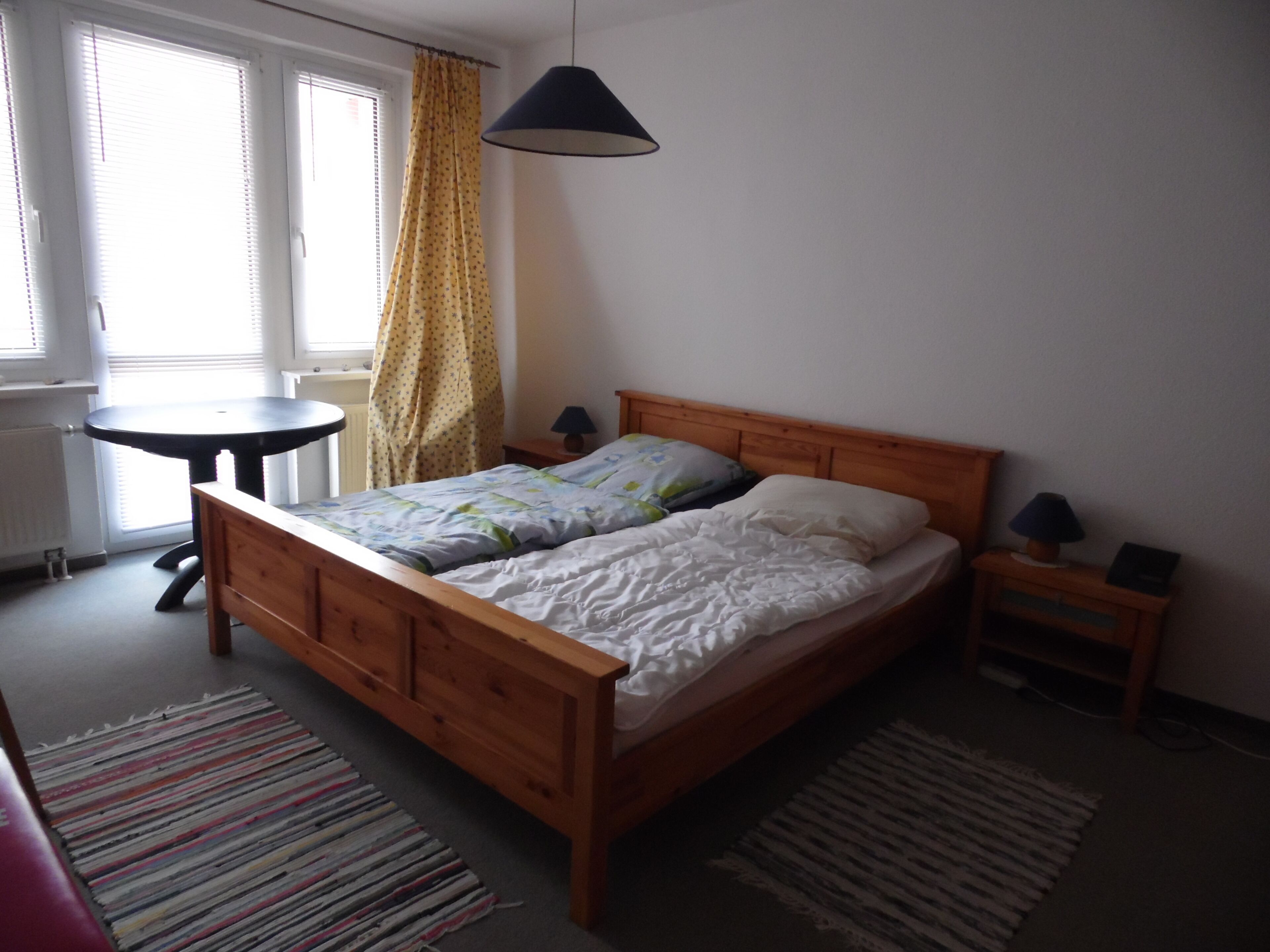 A bedroom with a wooden bed, a small bedside table, a lamp, a window with curtains, and a radiator.