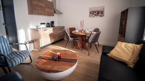 Ett modernt vardagsrum med ett runt soffbord, en soffa med en prydnadskudde, en matplats med stolar och ett bord samt ett köksområde med skåp och en kaffemaskin.