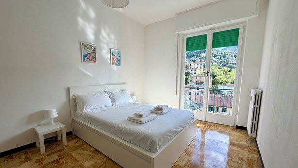 A bedroom with a bed, a nightstand, a lamp, a radiator, and a view of buildings through a window. - Schiappacasse 11 (Rapallo)