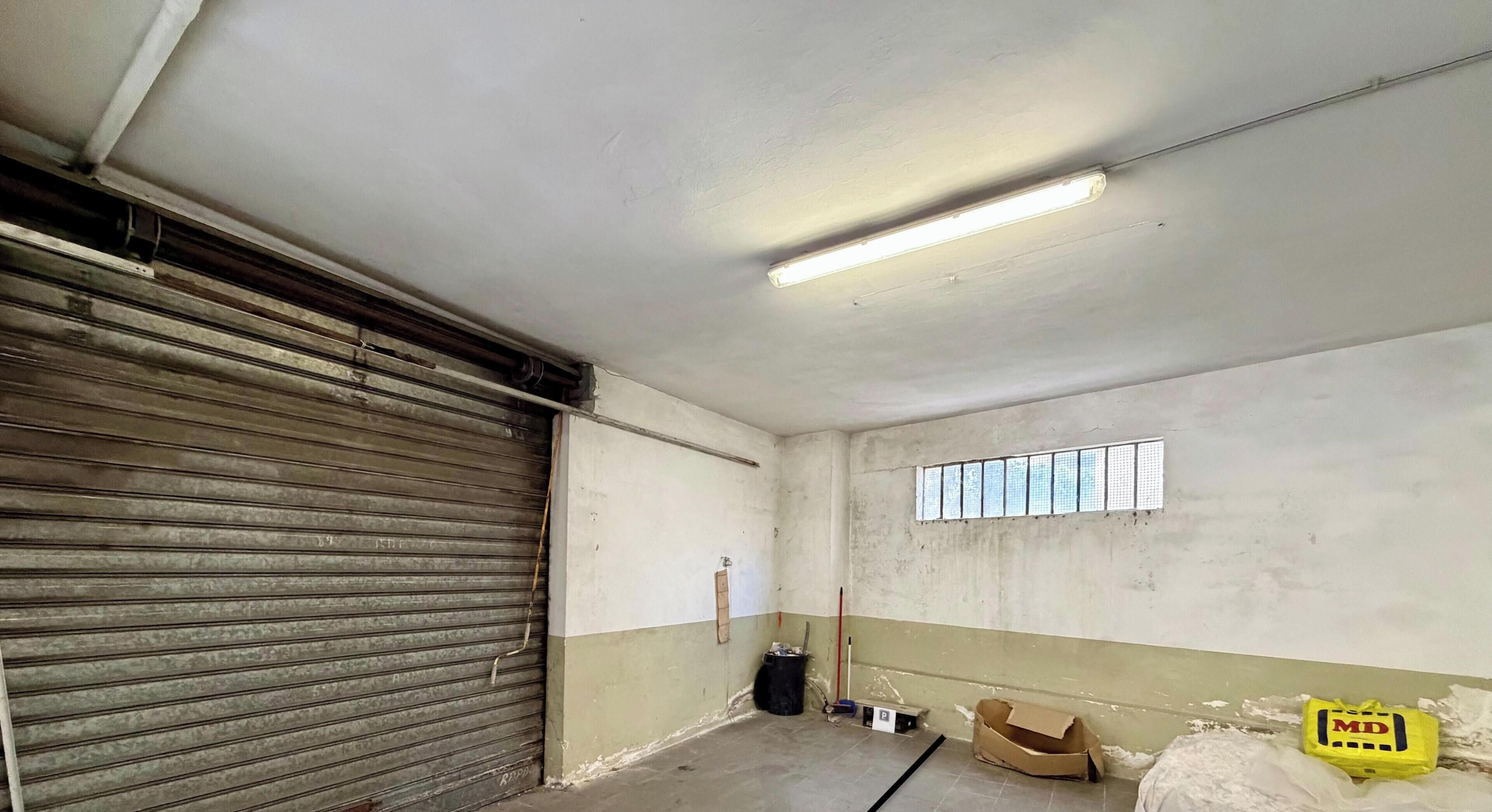 Une pièce avec une porte à volet métallique fermée, une fenêtre avec des stores, un luminaire fluorescent et une petite poubelle.