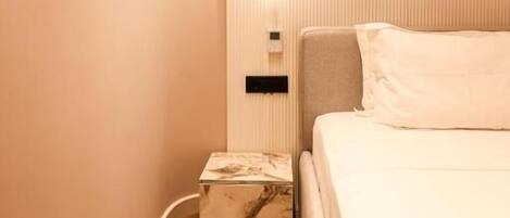 Una habitación de hotel con una cama, una mesita de noche, una lámpara fijada a la pared y una caja con estampado de mármol.