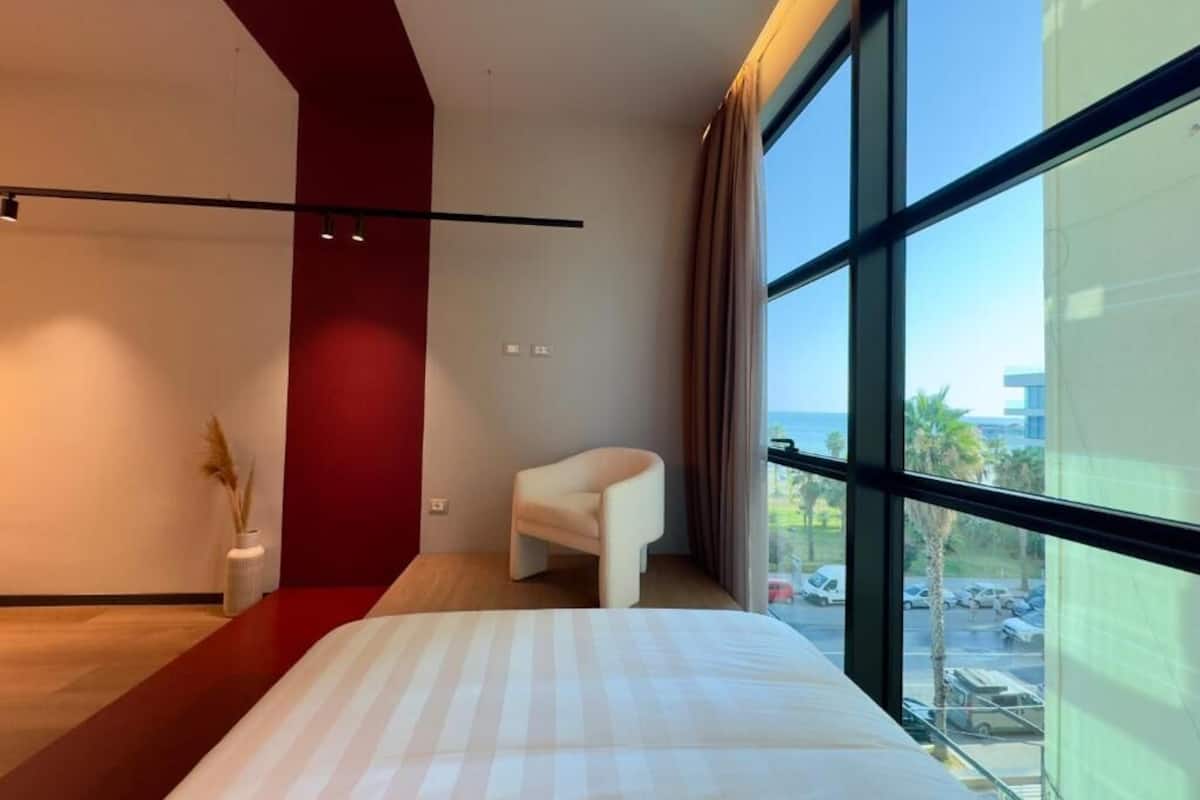 Una habitación de hotel moderna con una cama grande, una silla blanca, una pared de acento roja y vistas al estacionamiento a través de amplios ventanales.