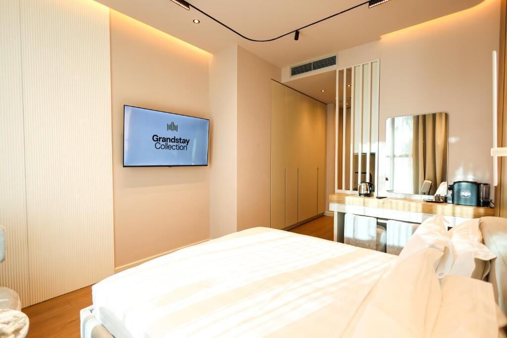 A hotel room with a bed, a television displaying 'Grandstay Collection,' a mirror, and a coffee machine.