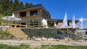 En moderne bygning med to etager, en træterrasse, en restaurant kaldet 'Pizzeria' og et anlagt område med beplantning.