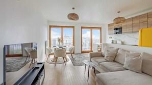 Ein modernes Wohnzimmer mit Fernseher, Sofa, Esstisch, Stühlen und großen Fenstern.