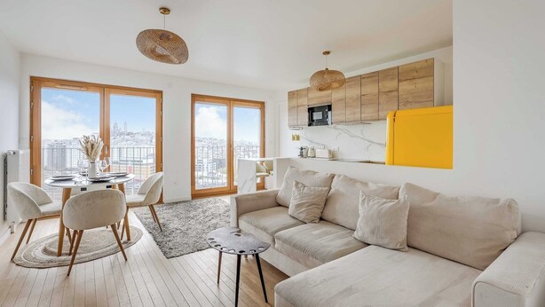 Ein modernes Wohnzimmer mit Sofa, Esstisch und Stühlen, eine Küche mit Holzausstattung und große Fenster mit Blick auf die Stadt.