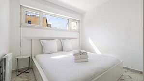 Une chambre moderne avec un grand lit, une petite table de chevet, un radiateur et une fenêtre donnant sur des immeubles.