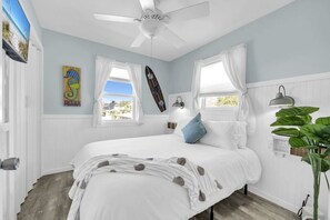A bedroom with a bed, two windows with curtains, a ceiling fan, a surfboard, a lamp, and a plant. - Sandy Toes Hideaway- Multi Family! (Panama City)
