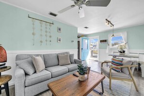 Un salon avec un canapé gris, une table basse en bois, un ventilateur de plafond et une vue sur l’extérieur, visible à travers une porte.