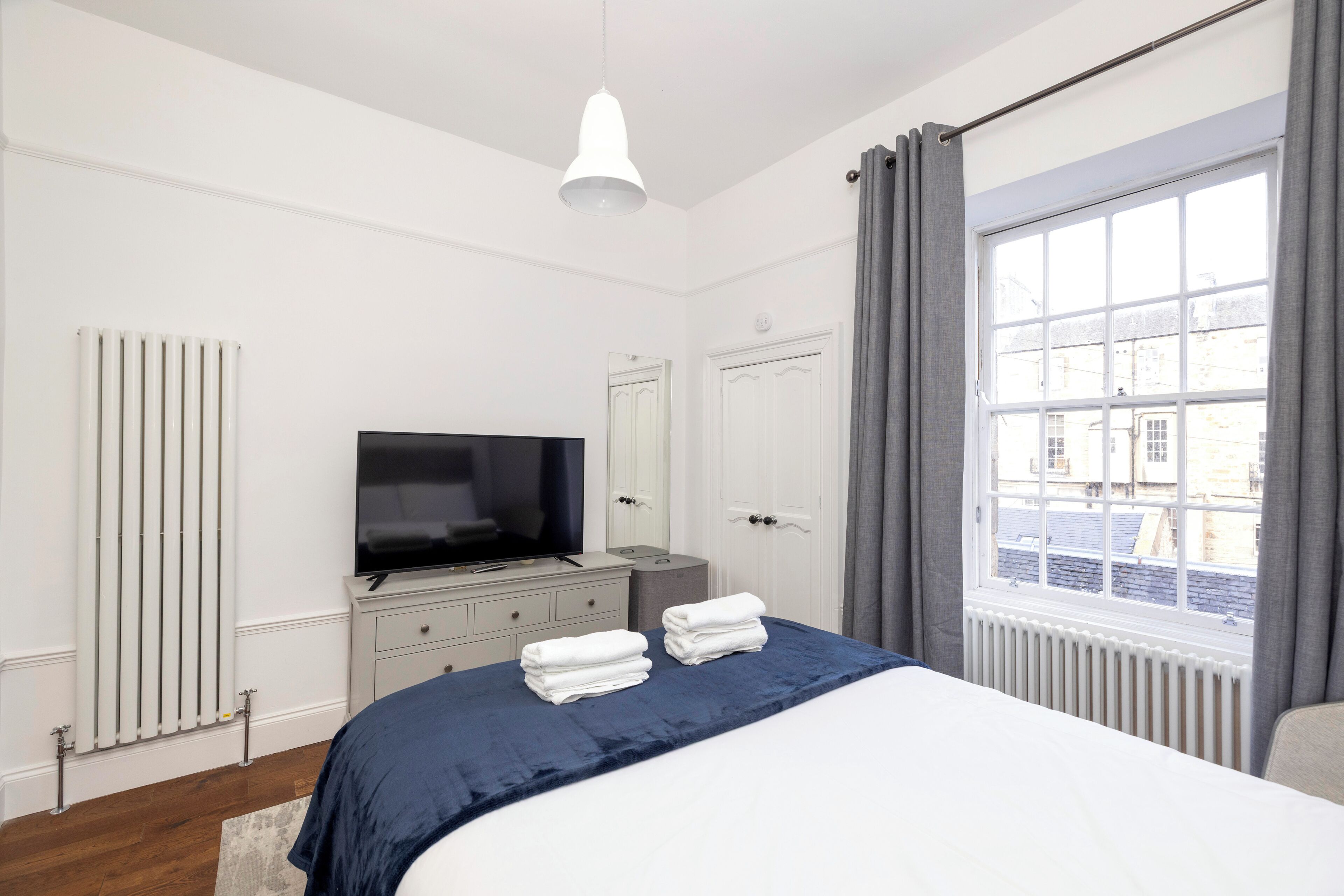 Un dormitorio con cama, televisor, cómoda, radiador y ventana con cortinas.