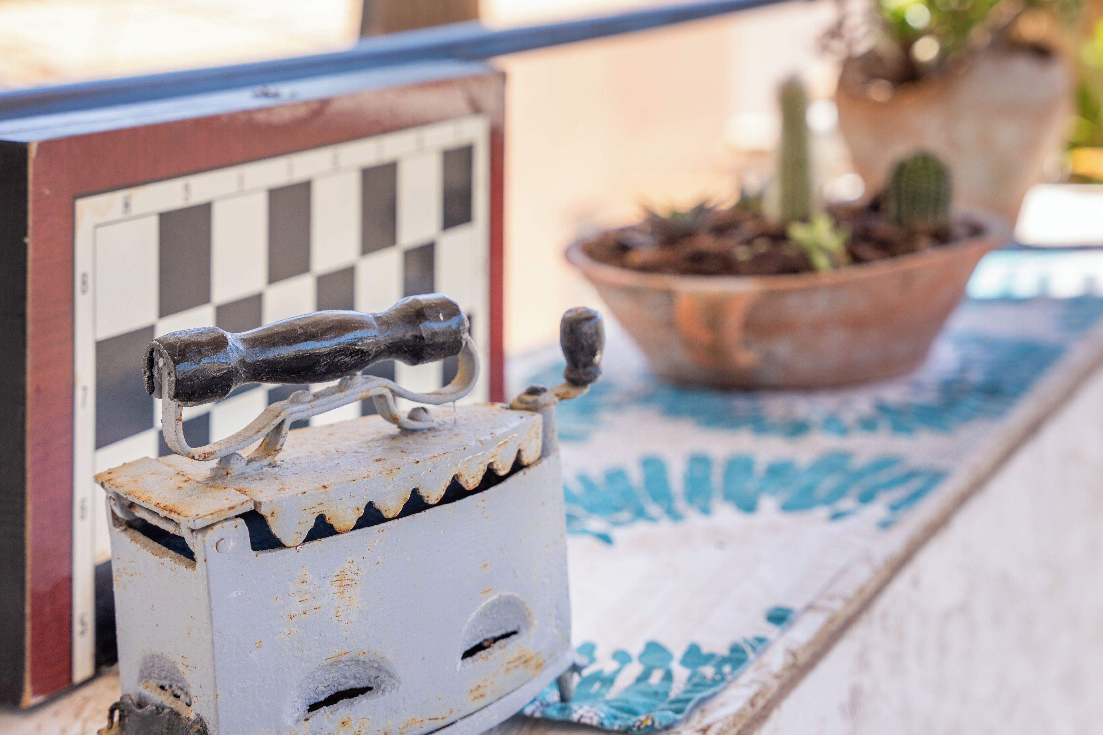 A rusty metal object with a handle, a checkered board, and a potted plant on a table.