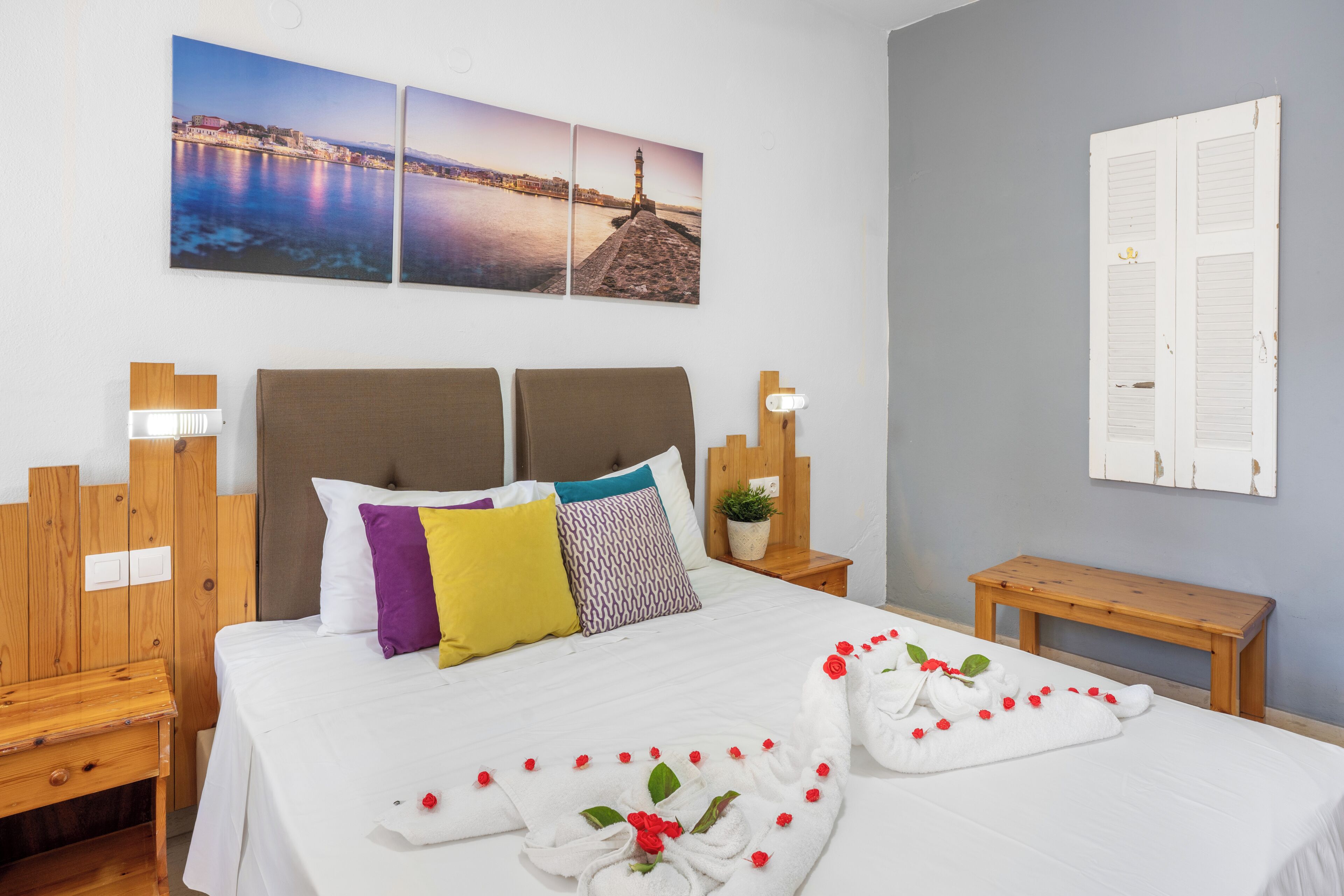 A hotel room with a bed, a nightstand, a bedside table, a wall-mounted lamp, and a picture of a coastal cityscape.