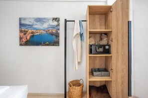 Ein Zimmer mit einem Bett, einem Holfach mit einer Mikrowelle, einem Korb und einem Gemälde einer Küstenstadt.