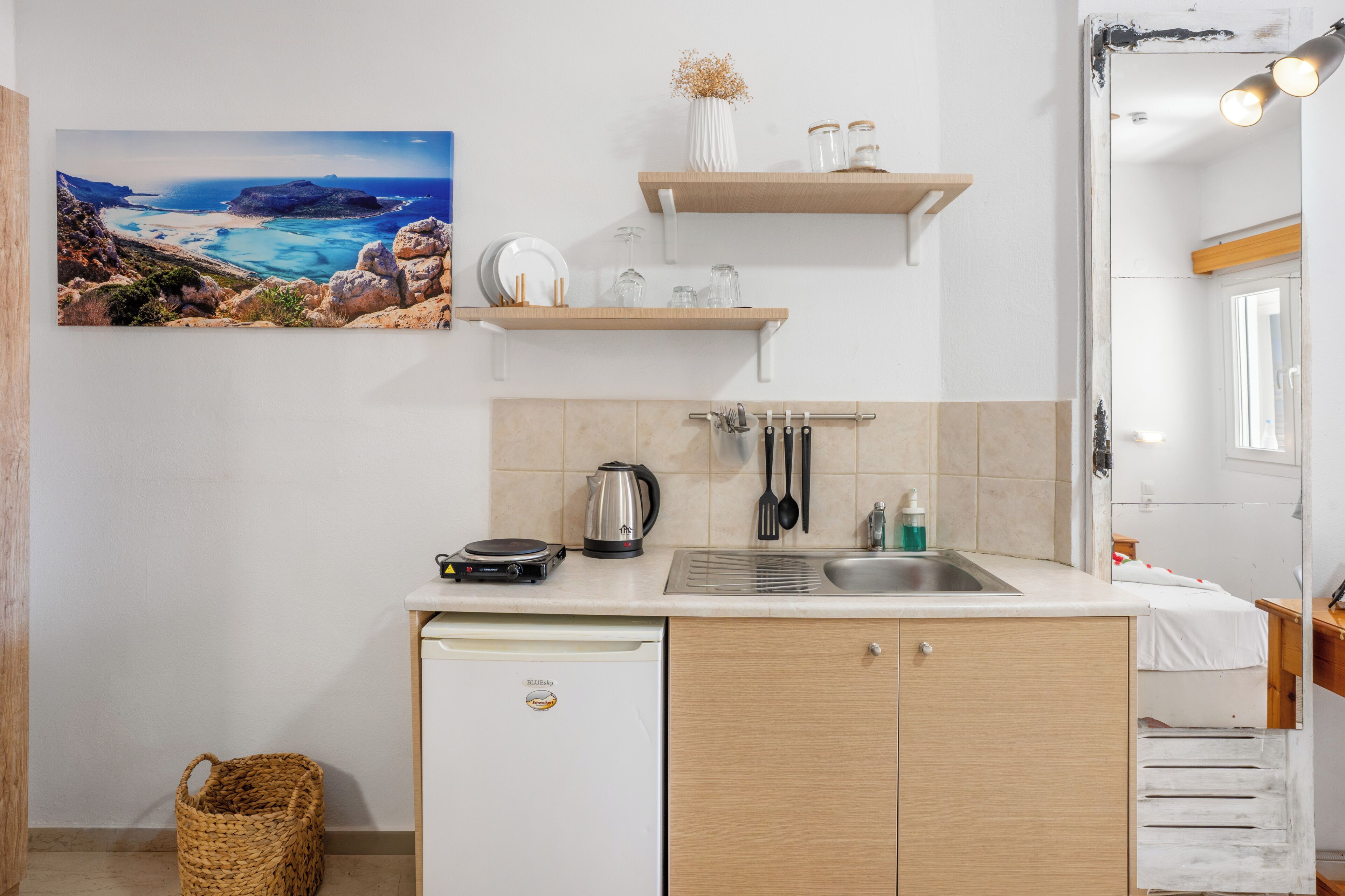 A compact kitchen with a sink, a coffee maker, and a small refrigerator. There are wooden shelves above the counter and a painting of a coastal landscape on the wall.