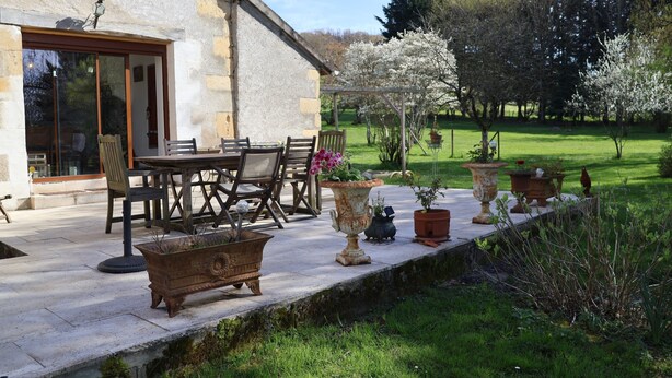 Une terrasse avec des meubles d’extérieur, un bâtiment en pierre et un jardin agrémenté d’arbres en fleurs.