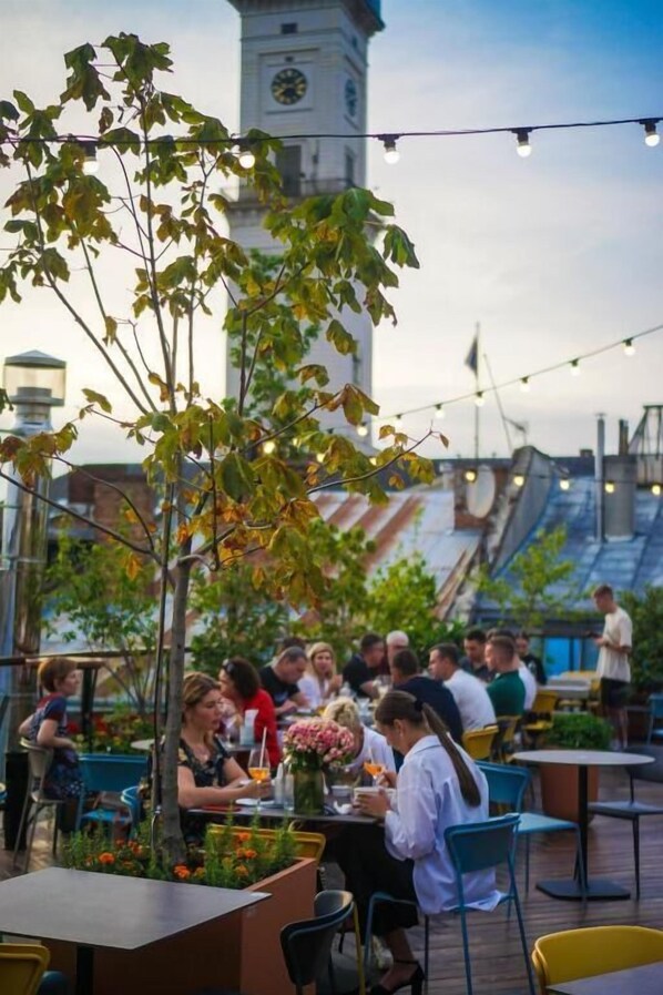 An outdoor dining area with tables and chairs, surrounded by greenery and a clock tower in the background. - FEST hotel. Rynok Square (Lviv)