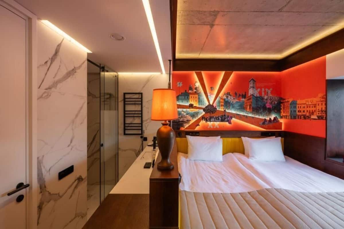 Une chambre d’hôtel comprenant un lit, une table de chevet, une lampe, une cabine de douche en verre et une fresque murale.