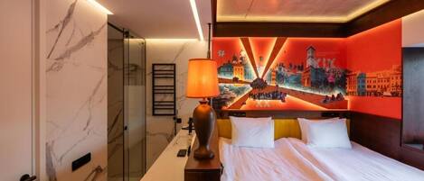 Una habitación de hotel con cama, mesita de noche, lámpara, cabina de ducha de vidrio y un mural en la pared.