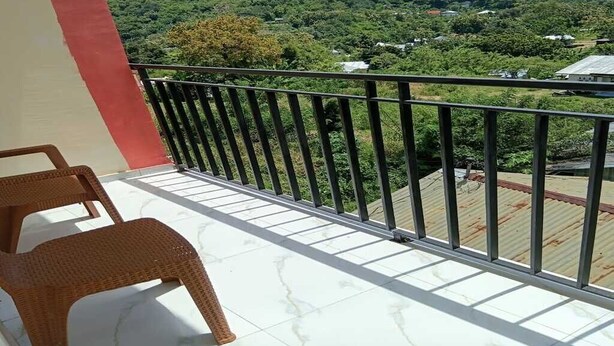 Een balkon met rieten stoelen, een metalen hekwerk en uitzicht op een groen landschap met bomen en gebouwen.