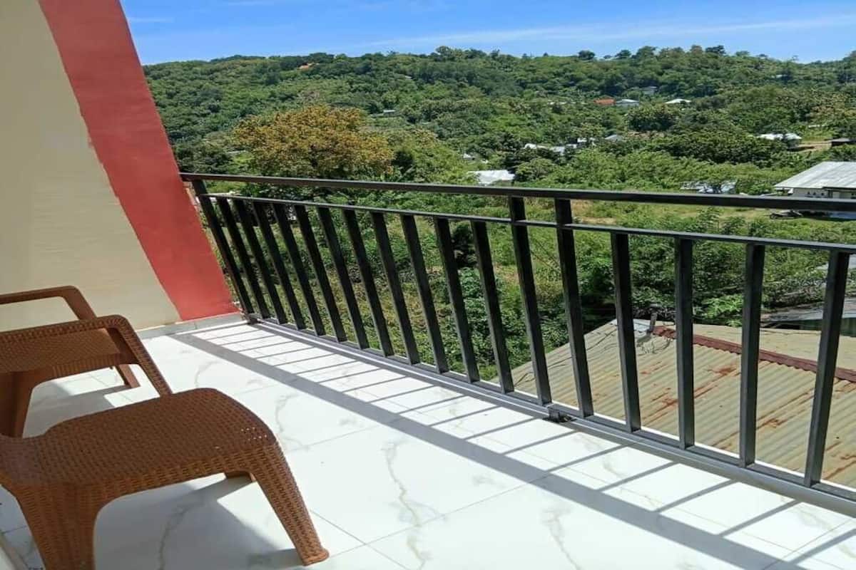 Ein Balkon mit Korbstühlen, einem Metallgeländer und Blick auf eine grüne Landschaft mit Bäumen und Gebäuden.