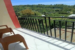 A balcony with wicker chairs, a metal railing, and a view of a green landscape with trees and buildings.