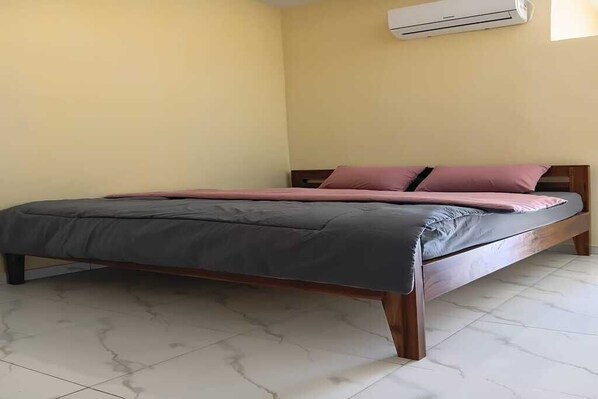 Un lit simple avec un cadre en bois, une couvre-lit grise et rose, et un appareil de climatisation blanc fixé au mur.