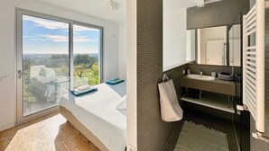 Une chambre dotée d’une grande fenêtre, d’un lit et offrant une vue sur un paysage. La salle de bain présente un design moderne avec un lavabo, un miroir et un porte-serviettes.