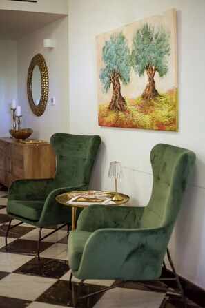 Ein Zimmer mit zwei grünen Sesseln, einem kleinen runden Tisch, einem Baumgemälde, einem Spiegel und einem Holzschrank.