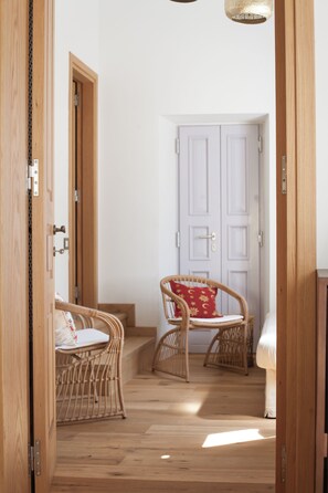A hallway with wooden flooring, wicker chairs, a white door, and a bed in the background. - Aylin Executive Suites - Saray Suite (Rhodes)