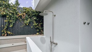 A modern outdoor shower with a metal hose and showerhead, adjacent to a white wall and a wooden deck. - Coastal Noir - Modern Cottesloe Stay (Cottesloe)