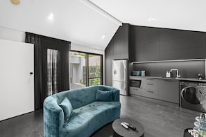 A modern kitchen with a built-in oven, microwave, and dishwasher, a blue sofa, and a large window. - Coastal Noir - Modern Cottesloe Stay (Cottesloe)