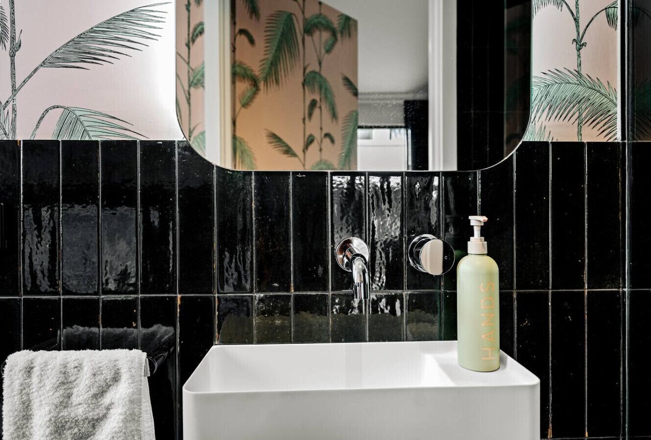 Une salle de bain avec un lavabo blanc, un miroir et un mur recouvert de carreaux noirs.