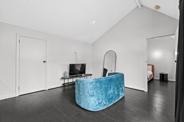 Un salon moderne avec un canapé bleu aux lignes courbes, un téléviseur à écran plat posé sur un meuble noir, un grand miroir et une porte donnant sur une autre pièce.