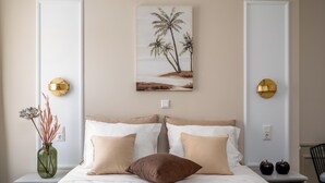 Een keurig opgemaakt bed met beige en bruine kussens, een groene vaas met gedroogde bloemen, en een schilderij van palmbomen aan de muur.