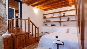 A bedroom with a wooden staircase, a bed with a white bedspread, a bedside table with a book and a glass of red wine, and a shelf with books and decorative items. - Belvedere Medieval Villa in Old Town (Old Town Rhodes)